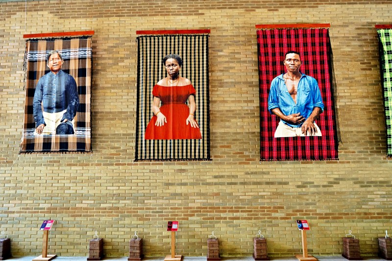 Três grandes painéis de tecido, representando figuras humanas, pendurados em uma parede de tijolos. A figura à esquerda está vestida com roupas tradicionais, a do meio usa um vestido vermelho e a da direita veste uma camisa azul. Abaixo dos painéis, há suportes com informações.