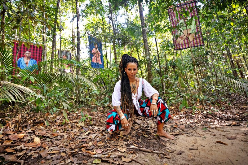 Mulher com cabelo longo e dreadlocks sentada no solo na floresta, vestindo roupas coloridas. Ao fundo, fotos em telas penduradas entre as árvores.