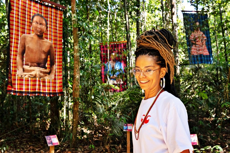 Mulher sorridente em um ambiente de floresta, com imagens penduradas ao fundo que representam culturas indígenas.