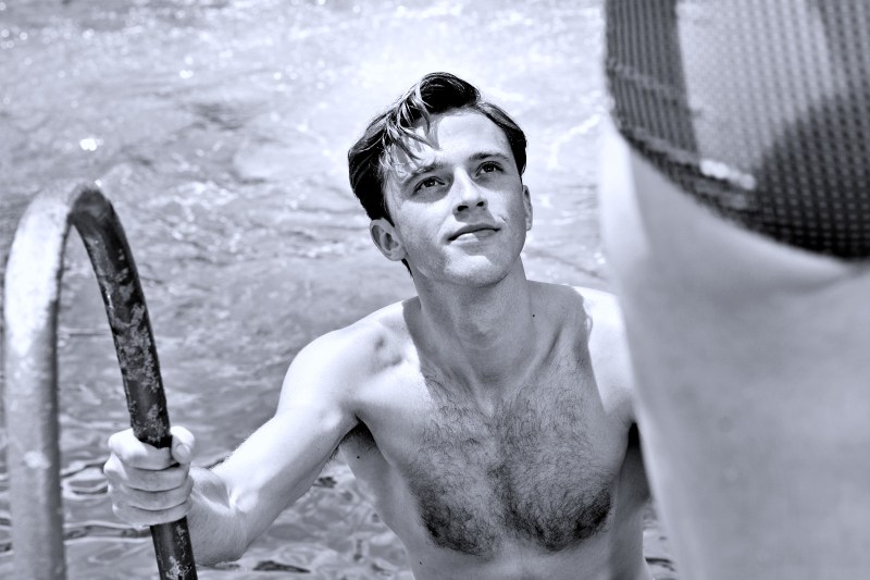 Um jovem sem camisa está subindo uma escada em uma piscina, olhando para cima com um sorriso, enquanto a parte inferior de uma pessoa de costas é visível ao lado.