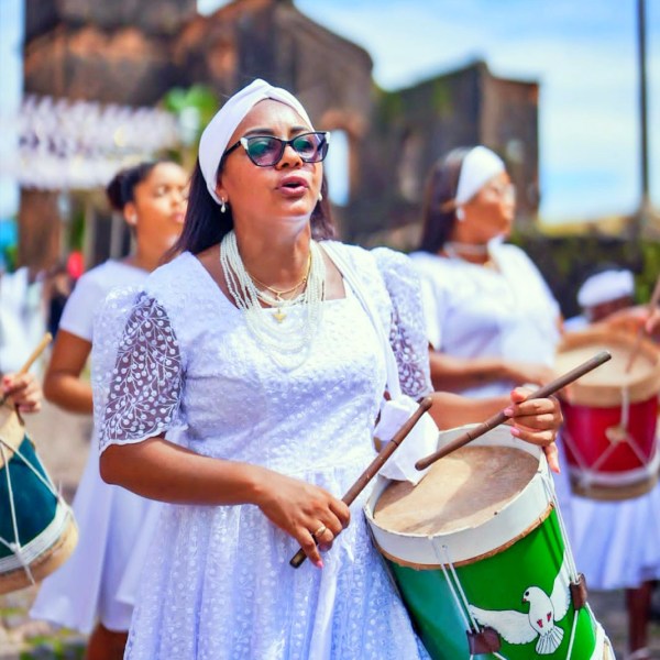 Mulher cantando e tocando tambor, vestida de branco, com óculos escuros. Outras mulheres em trajes semelhantes ao fundo, durante uma celebração ao ar livre.