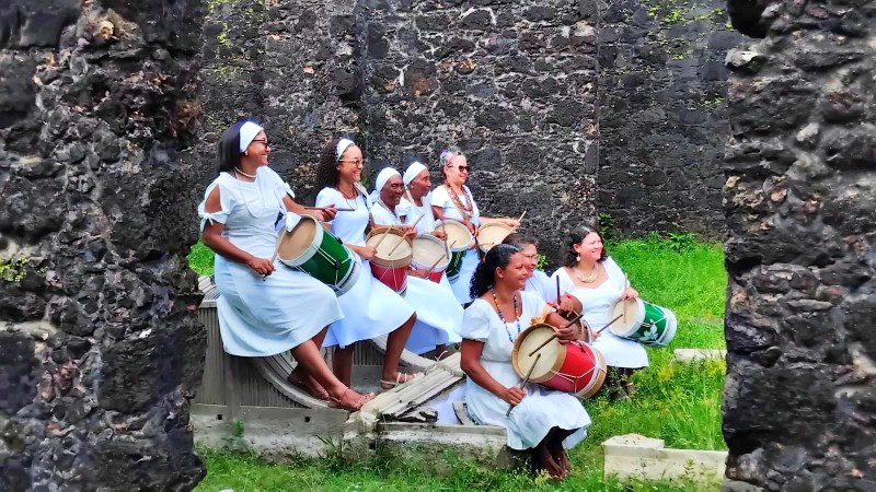 Grupo de mulheres vestindo roupas brancas, tocando tambores em um ambiente ao ar livre, entre ruínas de pedras.