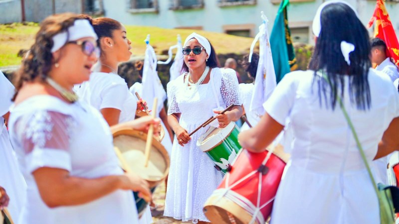 Grupo de mulheres vestidas de branco tocando instrumentos musicais e cantando durante uma celebração cultural.
