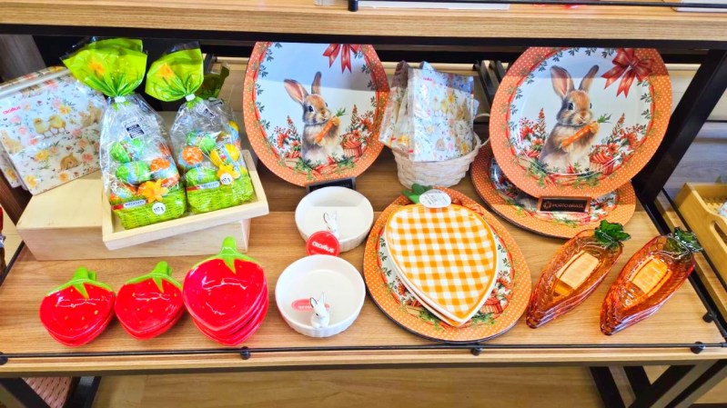 Conjunto de pratos decorativos com coelho, utensílios de cerâmica em forma de morango, e doces em embalagens coloridas em uma prateleira de madeira.