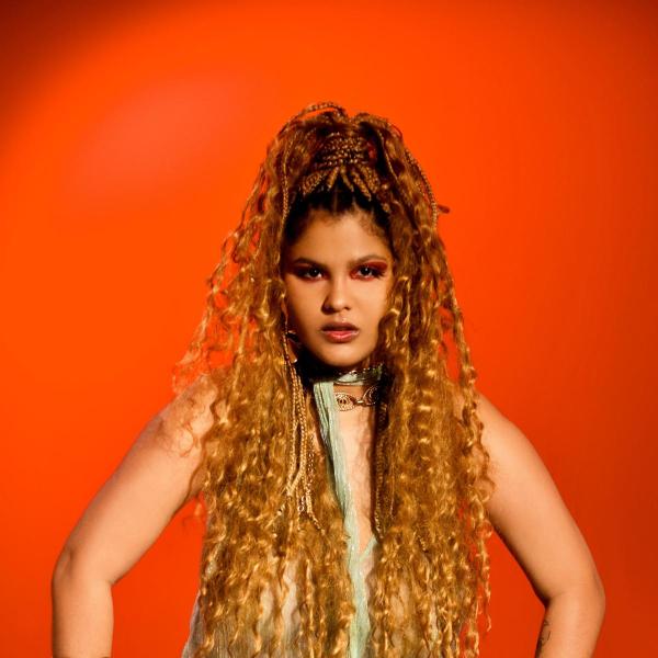 Pessoa com cabelo longo e cacheado, usando um vestido leve, contra um fundo laranja.