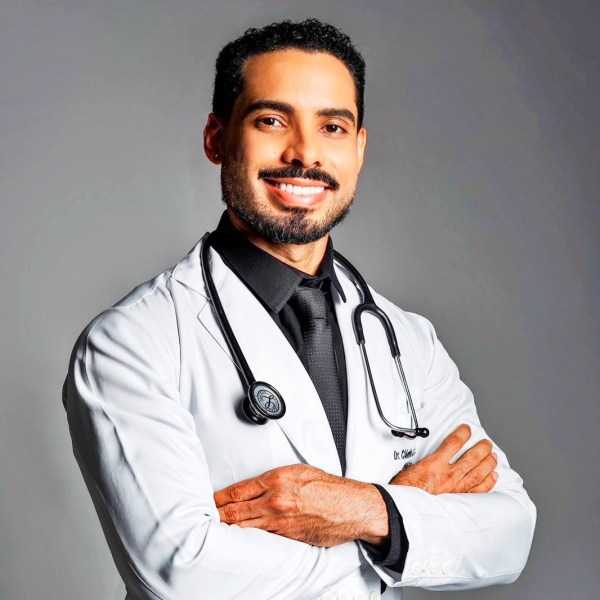 Homem sorridente vestindo jaleco branco com estetoscópio, posando com os braços cruzados.