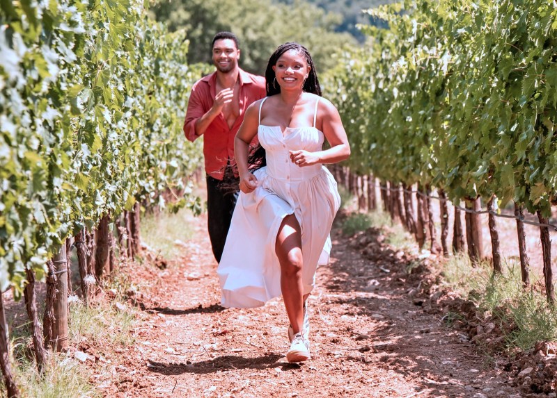 Casal correndo em uma vinícola, com parreiras ao fundo. A mulher está usando um vestido branco e o homem uma camisa vermelha.