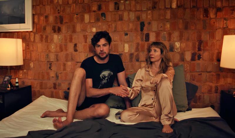 Casal sentado na cama, com o homem usando uma camiseta preta e a mulher vestindo um pijama claro. Eles parecem estar conversando ou confortando um ao outro em um ambiente acolhedor com parede de tijolos expostos.