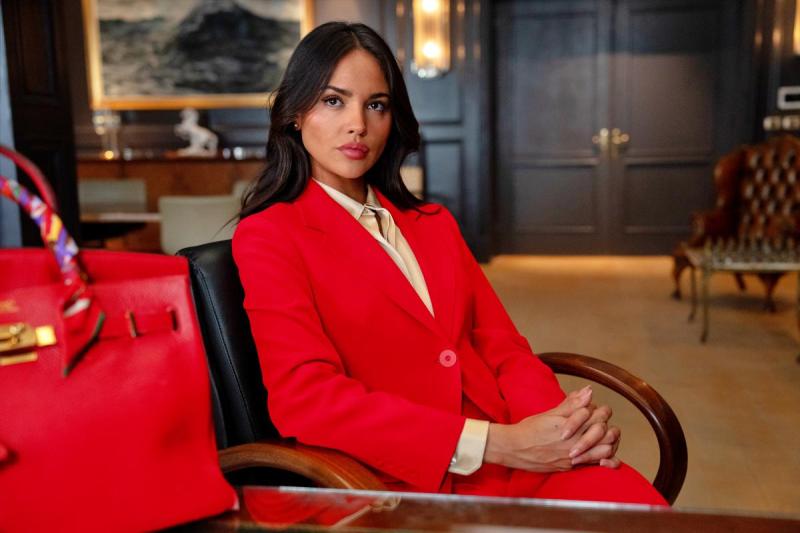 Mulher sentada em uma cadeira, usando um terno vermelho e uma camisa clara, com expressão confiante em um ambiente de escritório elegante, ao lado de uma bolsa vermelha.