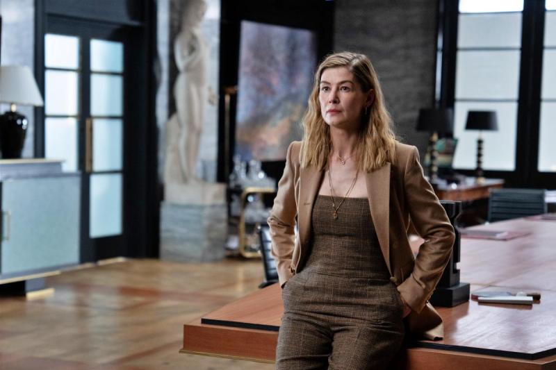 Mulher de cabelo loiro e ondulado, vestindo um conjunto marrom, em pé ao lado de uma mesa em um escritório moderno, com uma escultura e arte ao fundo.