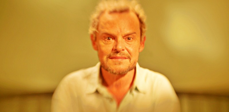 Retrato de um homem sorrindo com cabelo grisalho e barba, em um ambiente com iluminação suave e fundo desfocado.